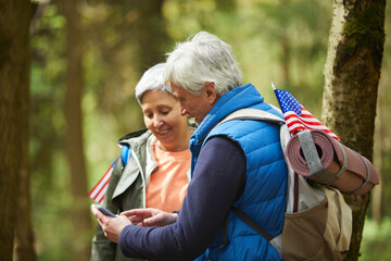 Side view at active senior couple using mobile app via smartphone while enjoying hike in forest, copy space