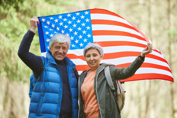 Waist up portrait of active senior couple holding American flag and smiling at camera while enjoying hike in forest, copy space