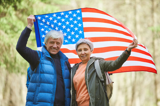 Waist up portrait of active senior couple holding American flag and smiling at camera while enjoying hike in forest, copy space