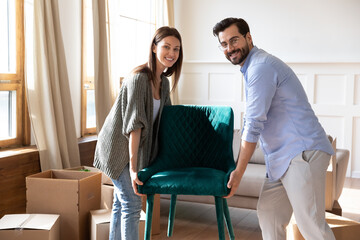 Portrait happy young couple carrying chair, placing furniture in living room in new house, looking at camera, moving day and renovation concept, excited family relocating into new apartment