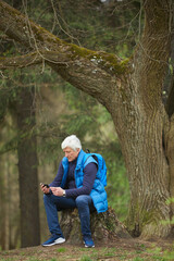 Fototapeta premium Vertical full length portrait of active senior man using smartphone while resting during hike in autumn forest, copy space above
