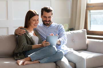Happy young couple using computer tablet together, sitting on cozy couch at home, smiling beautiful woman and man looking at screen, surfing internet, chatting online in social network