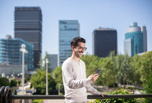 Joven trabajador con bigote y gafas sonriente mirando el tel&eacute;fono m&oacute;vil con un fondo de rascacielos a su espalda