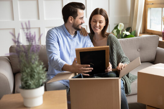 Happy wife and husband looking at photo frame, sharing memories, sitting on couch in living room, young couple unpacking cardboard boxes with belongings on moving day, relocation and mortgage