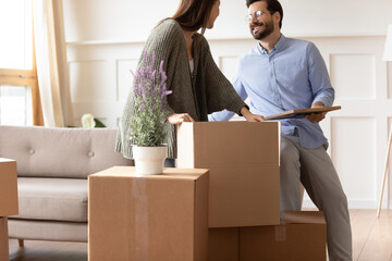 Excited young couple unpacking cardboard boxes in modern living room, moving day and mortgage concept, happy wife and husband preparing to relocation into new house, holding belongings