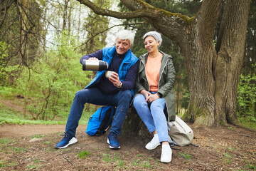 Full length portrait of cheerful senior couple enjoying hot tea from flask while resting during hike in autumn forest, copy space