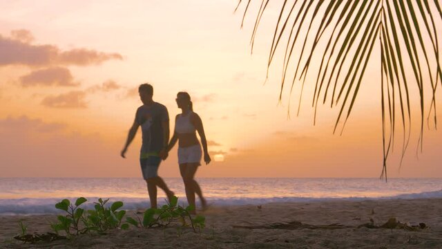 LOW ANGLE, DOF: Caucasian tourist couple holds hands while walking down the tropical sandy beach in the Caribbean at sunset. Young man and woman on summer vacation go for a stroll on a sunny evening.