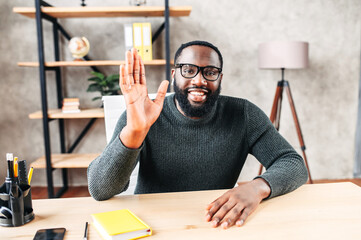 Webcam view of cheerful African-American young guy in glasses, he looks into camera and greeting , waving hello. Video screen, video chat, online call