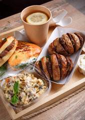 Disposable tableware, lemon tea, slate, mouth-watering sausage rolls and dill and baked potatoes with meat on a wooden tray. Side view.