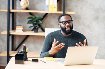 Video call. Attractive African-American guy is using laptop for video communication, a guy in glasses sits at the table in modern office, looking into webcam and talking online - Powered by Adobe