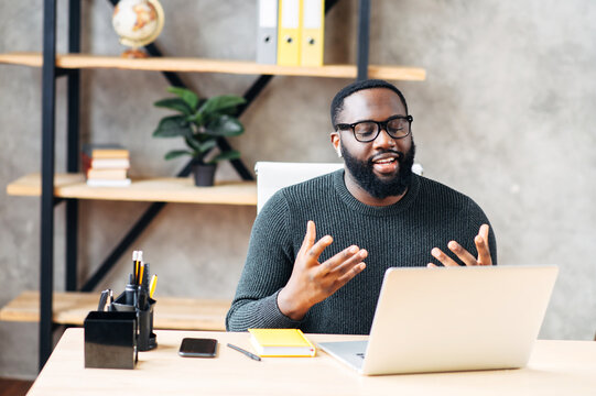 Video Call. Attractive African-American Guy Is Using Laptop For Video Communication, A Guy In Glasses Sits At The Table In Modern Office, Looking Into Webcam And Talking Online