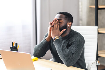 Side view of exhausted African-American office employee sits at the desk with a laptop and covers...