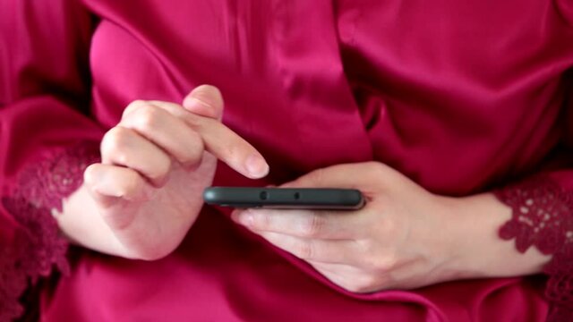 Woman using smartphone, girl in red lace nightie scrolling the screen of mobile phone by finger. Concept of online addiction, sms, social media