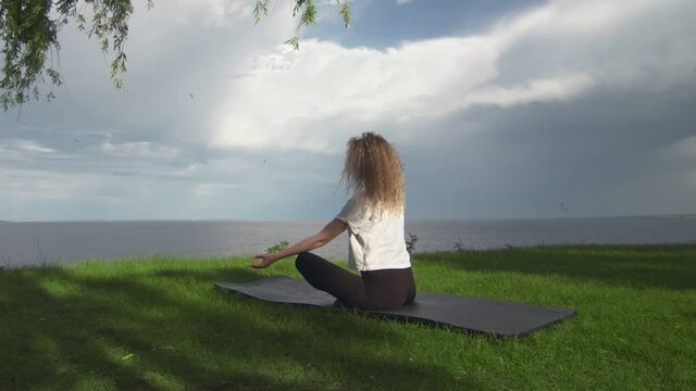 Back view of young fit woman practice yoga on coast near the lake or sea. Woman sitting in lotus pose and meditating