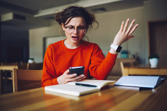 Angry attractive female feeling disappointed receiving bill from banking service checking email box in smartphone connected to wireless internet, emotional hipster girl upset about failure of payment