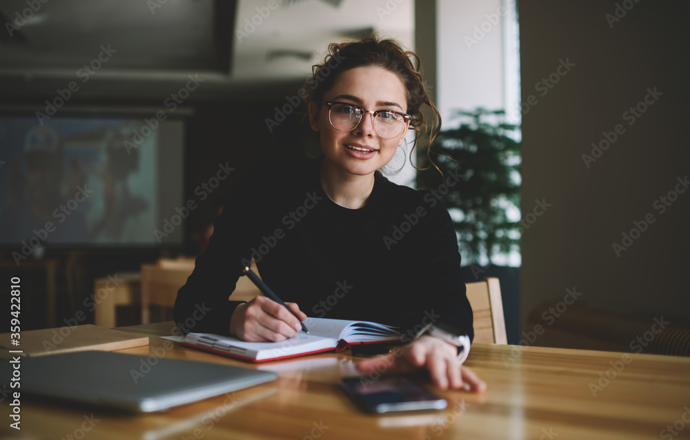 Fototapete portrait of intelligent student with optical spectacles writing some records in textbook while sitti