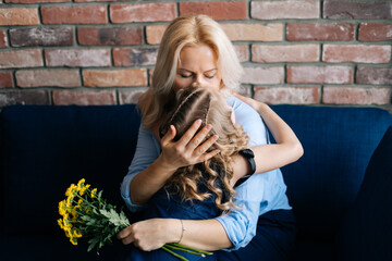 Mom kisses and hugs her little daughter, who gave her a bouquet of flowers at the home in cozy living room. Concept of Happy Mothers Day.