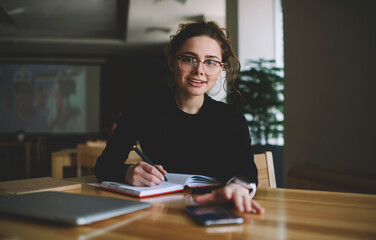 Portrait of intelligent student with optical spectacles writing some records in textbook while...