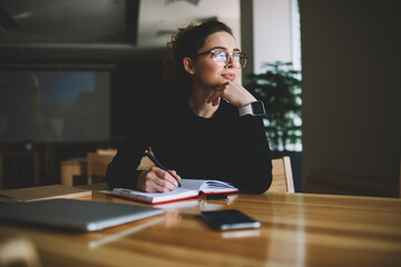 Thoughtful young author businesswoman and thinking on writing interesting article.Woman in stylish eyeglasses with good lenses looking out of window and pondering new plans while noting them - obrazy, fototapety, plakaty