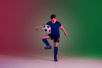 Fototapeta na wymiar Childhood. Teen male football or soccer player on gradient background in neon light. Caucasian boy training, practicing on the run, in jump. Concept of sport, competition, winning, motion, action.