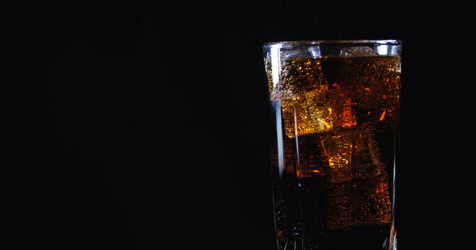Cola with ice cubes. Side view of rotating glass in which cold carbonated brown sweet drink is poured on black background isolated with free copy space. Slow Motion 50 FPS 4k