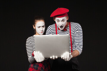 man and woman with white makeup on their faces like mimes looks at a laptop on a black background