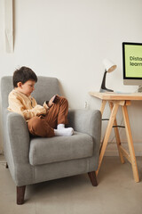 Vertical full length portrait of cute boy sitting cross-legged in armchair and using smartphone, copy space