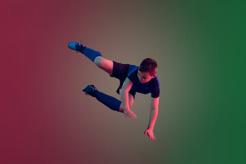 Unstoppable. Teen male football or soccer player on gradient background in neon light. Caucasian boy training, practicing on the run, in jump. Concept of sport, competition, winning, motion, action.