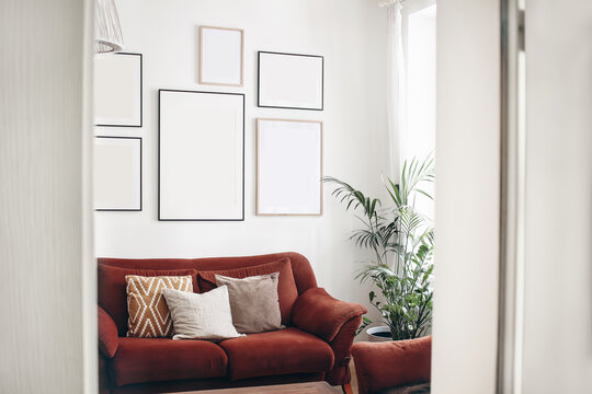 Blank picture frames mockups on white wall. White living room design. View of modern boho, scandi style interior with sofa, cushions, potted palm plant through open white door. Home staging concept.