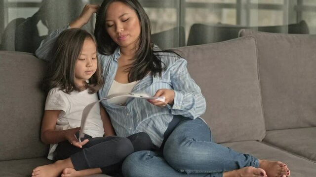 A happy young asian mother is reading a book with her daughter while sitting in the living room at home