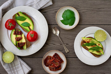 Bruschetta style rye bread toasts with avocado, sun-dried tomatoes, basil, dill and lime. Rustic dark wooden background