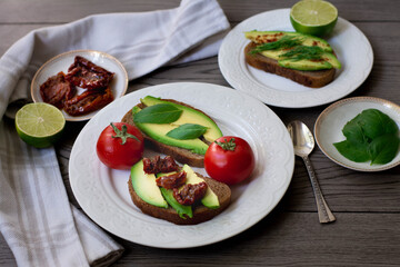 Bruschetta style rye bread toasts with avocado, sun-dried tomatoes, basil, dill and lime. Rustic dark wooden background