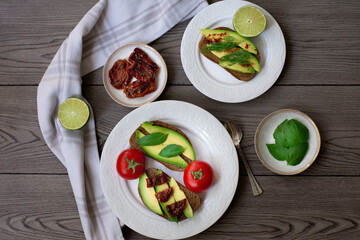 Bruschetta style rye bread toasts with avocado, sun-dried tomatoes, basil, dill and lime. Rustic dark wooden background