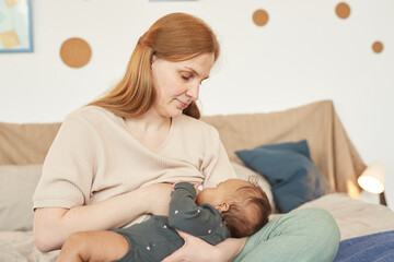 Warm-toned portrait of happy adult mother breastfeeding mixed-race baby while sitting on bed at...