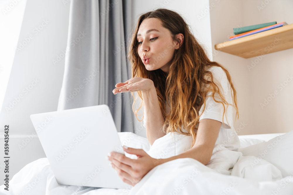 Poster portrait of beautiful woman blowing air kiss and using laptop in bed