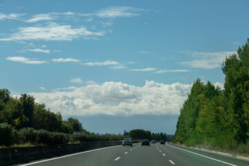 The highway is surrounded by green trees, blue sky and cumulus clouds ahead