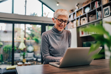 Portrait of a cheerful mature businesswoman working on laptop at home office. - obrazy, fototapety, plakaty