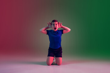 Winning, goal, screaming. Teen male football player on gradient background in neon light. Caucasian boy training, practicing on the run, in jump. Concept of sport, competition, winning, motion, action