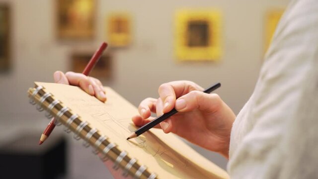 young female artist draws simple sketches in paper notebook with brown pages using black pencil in brightly lit hall of old art museum extreme close view