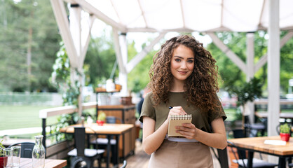 Waitress with order pad standing outdoors on terrace restaurant, looking at camera.