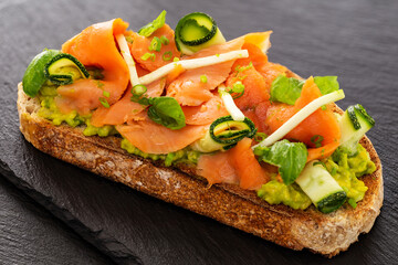 Open faced sandwich or French tartine with smoked salmon, avocado, zucchini rolls, basil and minced chives and apple on a toasted slice of sourdough bread. Displayed on slates.