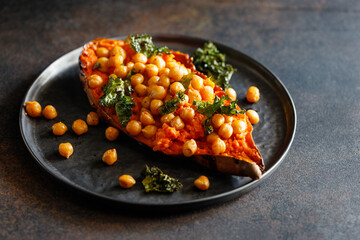 Baked sweet potato stuffed with chickpea and crunchy kale on a black plate. Vegan tasty dish for dinner or lunch.