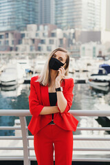 Obraz premium Young businesswoman in red suit and protective face mask standing at city promenade and making a call via a smarphone
