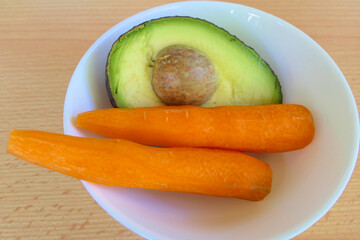 Healthy juice ingredients - closeup half avocado and carrots in a white bowl on a table 