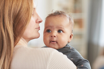Warm-toned close up portrait of cute mixed-race baby looking at camera while sitting in mothers arms at home, copy space