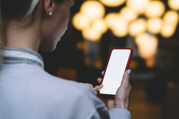 Back view from female shoulder of touching with finger lighted screen of smartphone device near copy space area with bokeh night lights. Texting message on mobile phone gadget with blank display