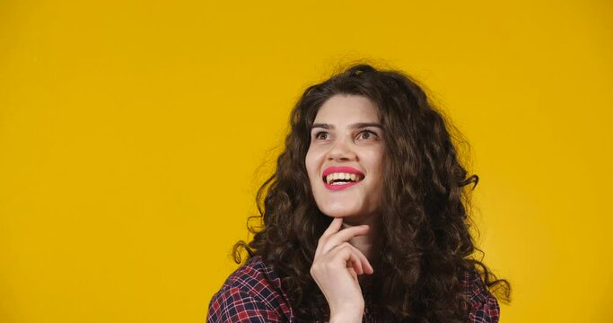 Curly-haired girl on a bright yellow studio background demonstrating different emotions. Looking thoughtful but then found  solution and joyfully pointing with her index finger