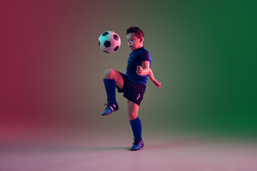 Fototapeta na wymiar Overcoming. Teen male football or soccer player on gradient background in neon light. Caucasian boy training, practicing on the run, in jump. Concept of sport, competition, winning, motion, action.