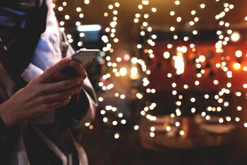 Closely image of young hipster girl is using her smart mobile phone, while is standing on the street against christmas garlands lights. Woman chatting with friends in social network via cell telephone