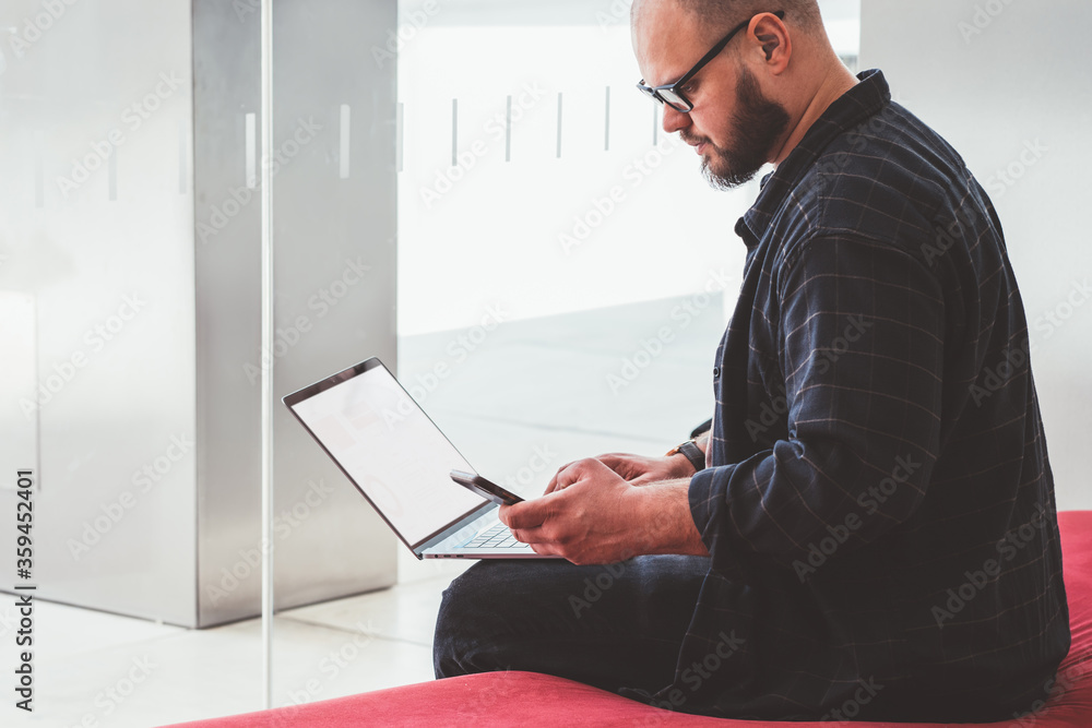 Poster pensive bearded young man reading notification on smartphone device while working remotely at laptop